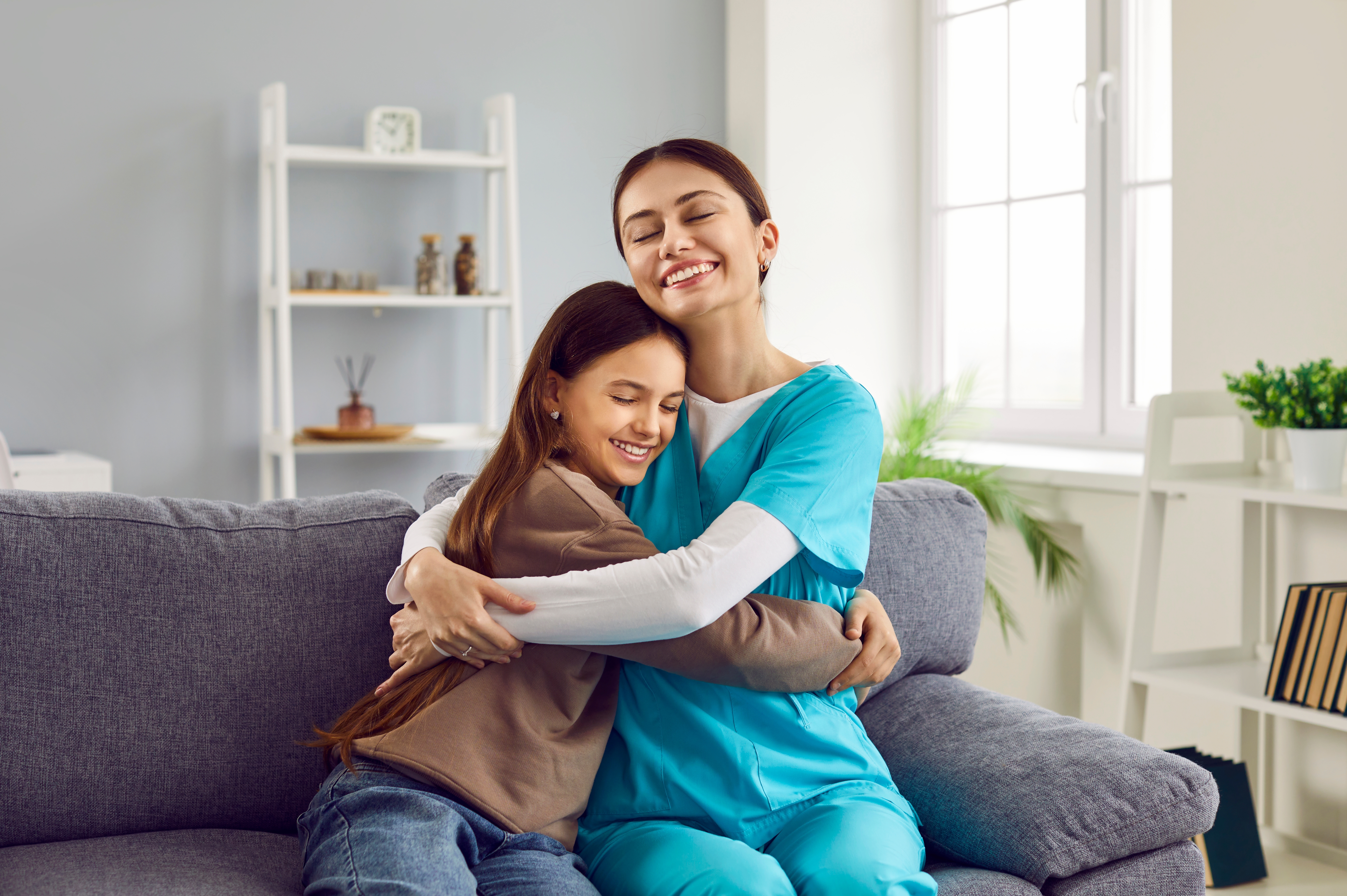Smiling Woman Pediatrician in Uniform and Little Girl Patient Sitting on Couch at Home and Hugging